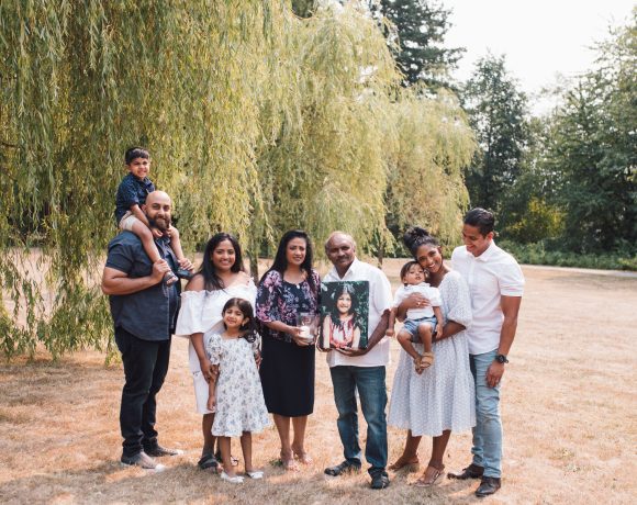 A family portrait outdoors with nine members of the Premia family across three generations, posing on a grassy field with a willow tree in the background. On the left, a man with a beard holds a young boy on his shoulders. Next to him, a woman in a white off-shoulder dress smiles beside a young girl in a floral dress. In the center, an older man and woman hold a framed photo of a teen girl, as a tribute. On the right, a woman holding a baby stands close to a man in a white shirt. They are all dressed in semi-formal attire, looking happy and connected.