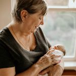 An affectionate moment between a grandmother and her grandchild. The elderly woman, with short grey hair and wearing a dark grey top, is holding and gazing lovingly at a newborn baby wrapped in a white blanket. The baby appears peaceful and content in her arms. They are indoors, near a window with a view of greenery outside, in a room filled with soft natural light.