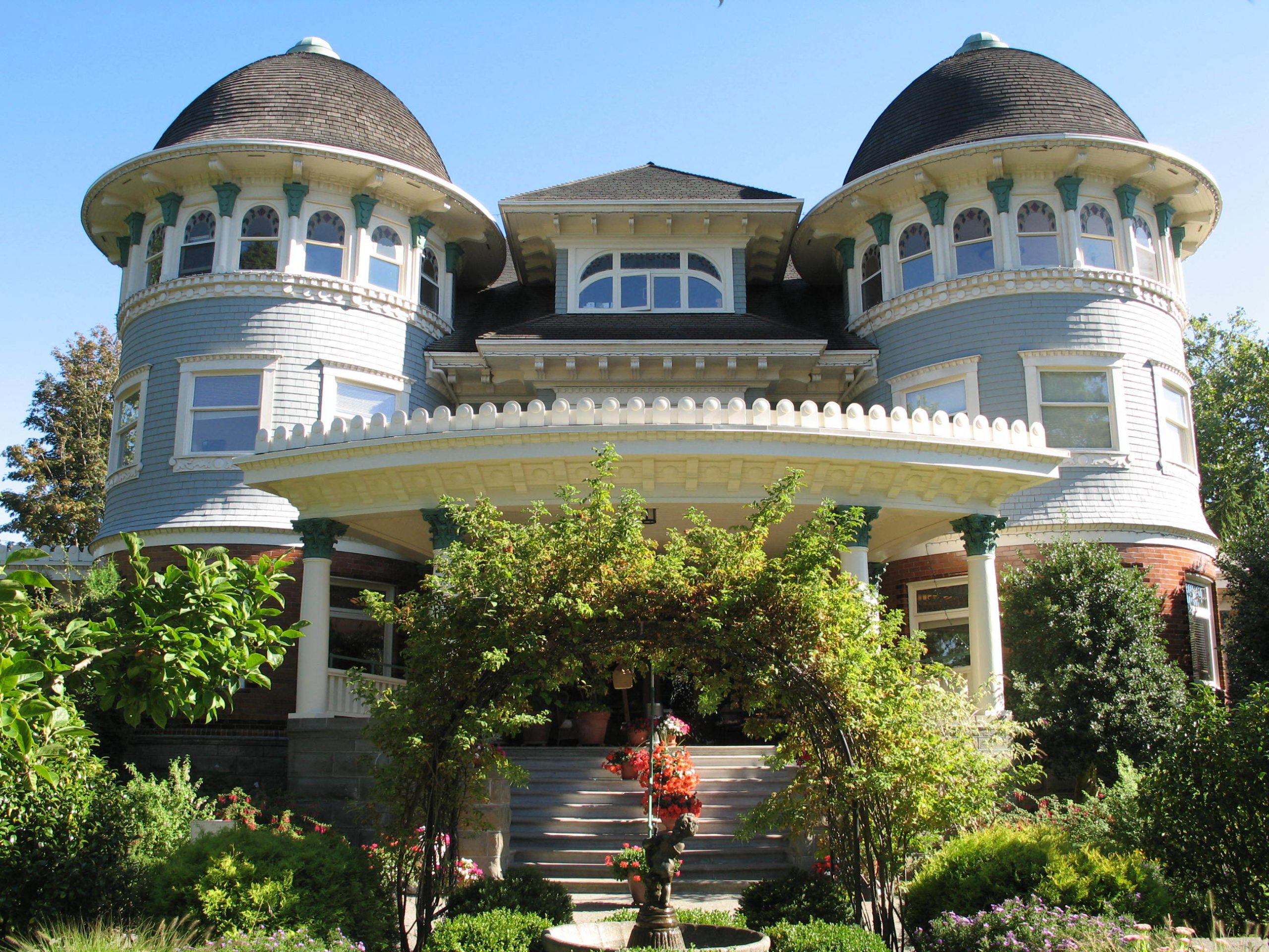 A vibrant summer view of the Canuck Place Vancouver mansion, a majestic Victorian-style house with distinctive twin towers and wrap-around porches. The house is painted in pale blue with white trim, featuring ornate detailing around the windows and roof edges. It is surrounded by a lush garden with a variety of plants and a well-kept lawn.