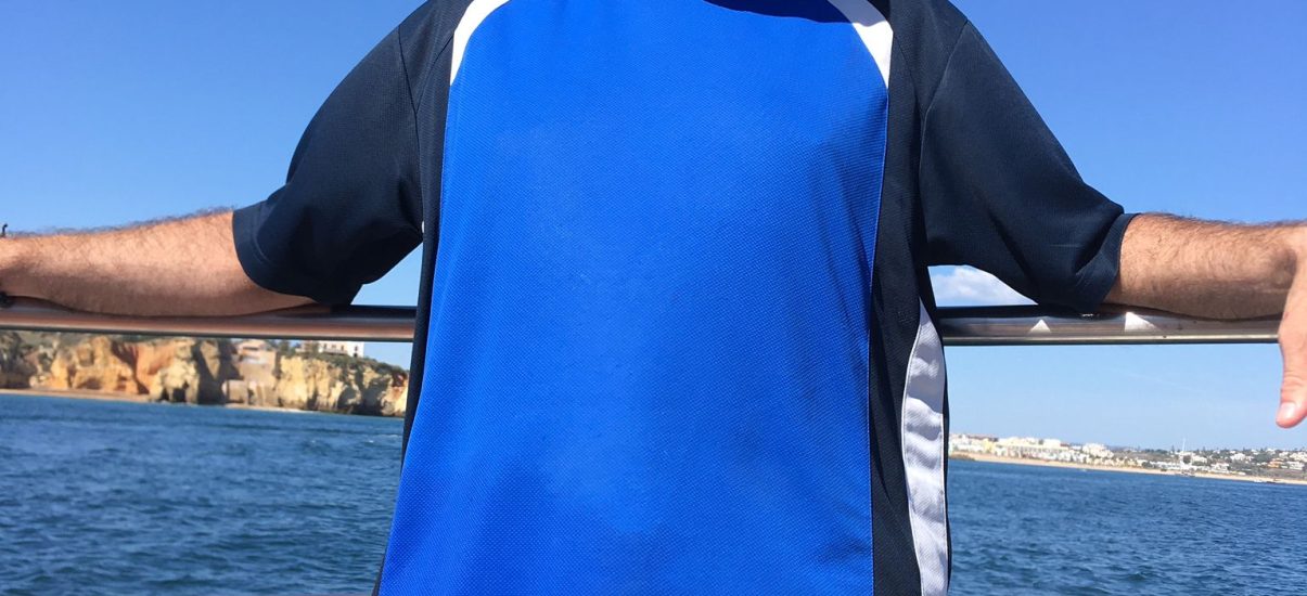 A middle-aged man in a bright blue t-shirt with dark blue sleeves and a light blue baseball cap and large black sunglasses is standing on a boat, smiling towards the camera.