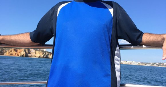 A middle-aged man in a bright blue t-shirt with dark blue sleeves and a light blue baseball cap and large black sunglasses is standing on a boat, smiling towards the camera.