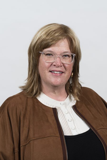 Professional headshot of a woman with blonde hair wearing clear glasses, a white button-up shirt, and a brown jacket, smiling against a plain white background.