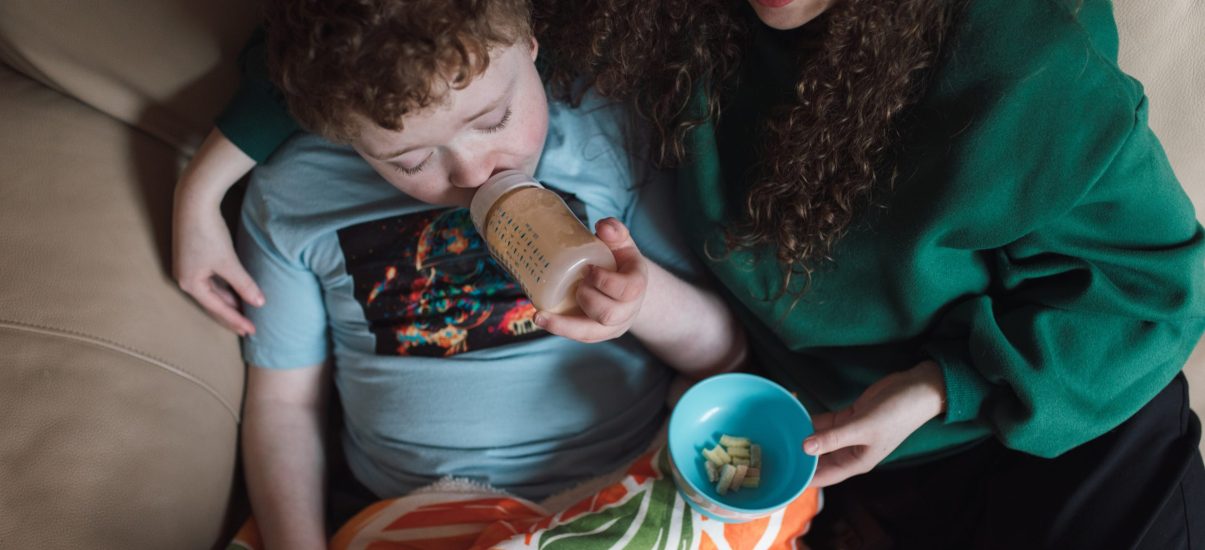 A mother sits on a couch with her young child, who is drinking from a bottle and watching something on an iPad. The mother is handing the child some food, creating a cozy and nurturing atmosphere as they share this moment together.