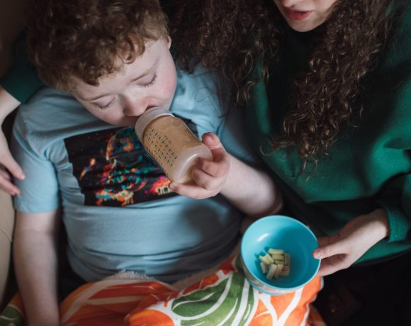 A mother sits on a couch with her young child, who is drinking from a bottle and watching something on an iPad. The mother is handing the child some food, creating a cozy and nurturing atmosphere as they share this moment together.