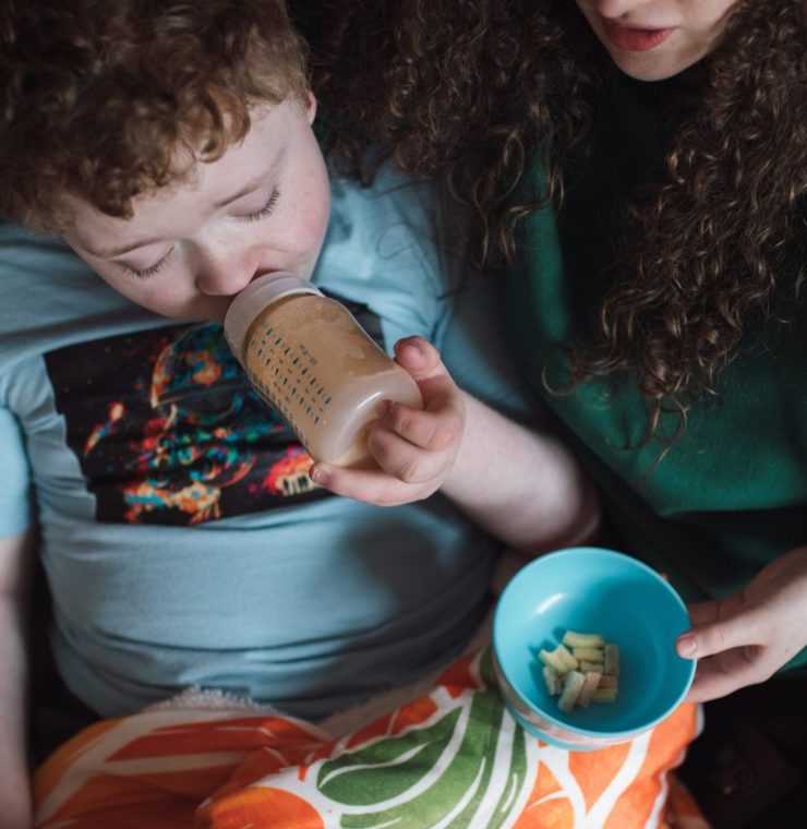 A mother sits on a couch with her young child, who is drinking from a bottle and watching something on an iPad. The mother is handing the child some food, creating a cozy and nurturing atmosphere as they share this moment together.