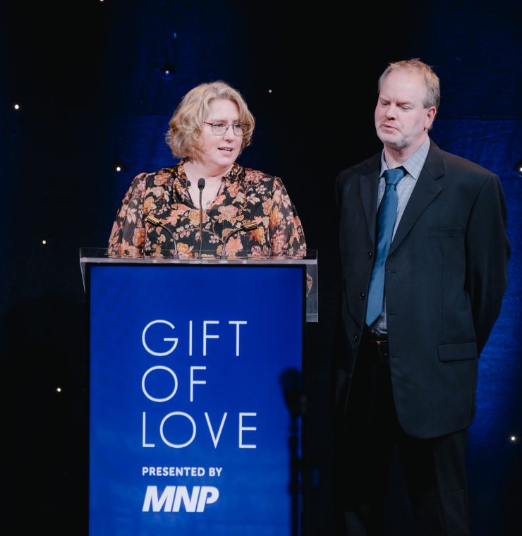 A white woman and a white man stand on a dimly lit stage behind a podium. The words ‘Gift of Love presented by MNP’ are written on the podium. The woman, with short blonde hair and glasses, wears a floral-patterned dress and speaks into the microphone with a confident expression. The man, dressed in a dark suit with a blue tie, stands beside her with his hands behind his back, listening with a solemn expression. The stage is illuminated with soft blue lighting, with small twinkling lights in the background adding a starry effect.