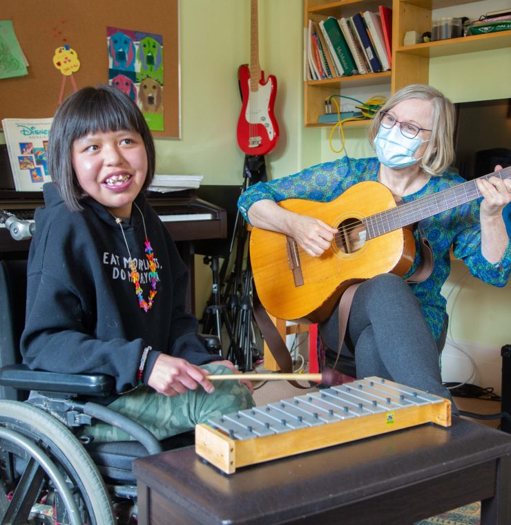 A young girl with shoulder-length dark hair and a joyful smile sits in a wheelchair, holding a small mallet and playing a wooden xylophone. She is wearing a black hoodie with colorful text and a beaded necklace. Beside her, an older white woman with short blonde hair, glasses, and a blue patterned blouse plays an acoustic guitar. She is wearing a light blue surgical mask. The setting is a music room with a piano, bookshelves filled with books and instruments, and a red electric guitar hanging on the wall. The two are engaged in a music therapy session.