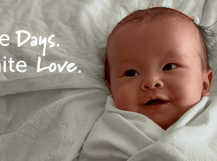 A baby of East-Asian descent with a small tuft of dark hair lies swaddled in a soft white blanket in a hospital crib. The baby has a gentle smile and wide, curious eyes looking to the side. A medical tube is visible in the background. The baby appears calm and comfortable, wrapped snugly in the blanket. The Canuck Place tagline