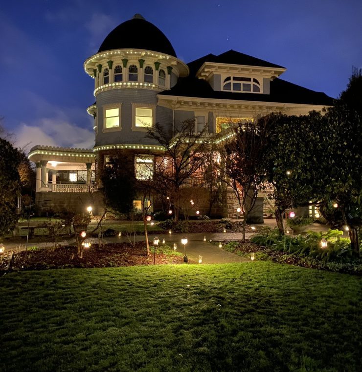 A heritage-style building at night, surrounded by a lush garden adorned with glowing lanterns. The warm lights outline the architectural details of the house to convey a welcoming and serene atmosphere. A pathway winds through the illuminated garden.