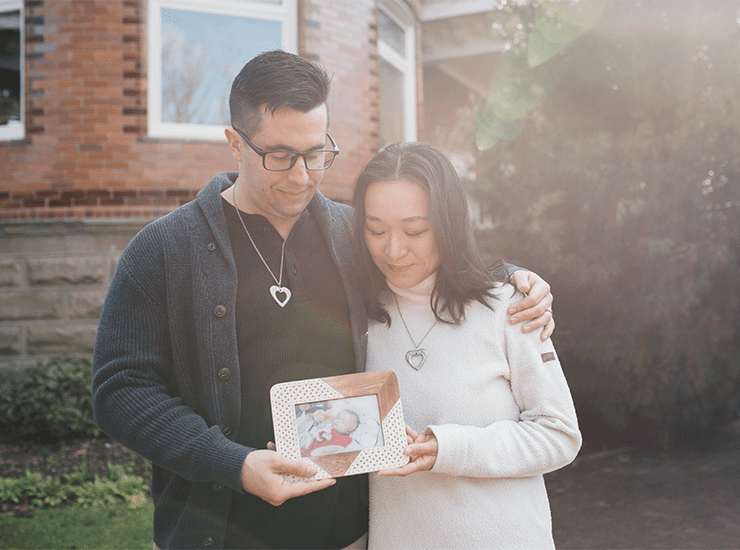 A couple stands together outside a brick building, bathed in soft sunlight. The man, wearing glasses and a dark sweater, gently wraps his arm around the woman, who is dressed in a light-colored sweater. Both wear matching heart-shaped necklaces. They hold a framed photograph of a baby, looking down at it with expressions of love and remembrance.