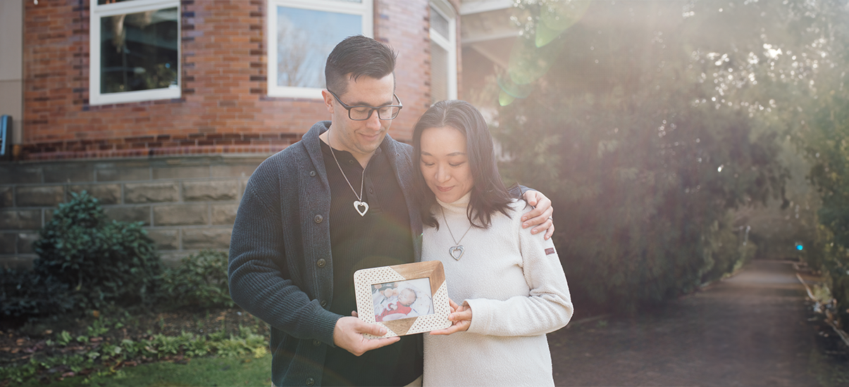 A couple stands together outside a brick building, bathed in soft sunlight. The man, wearing glasses and a dark sweater, gently wraps his arm around the woman, who is dressed in a light-colored sweater. Both wear matching heart-shaped necklaces. They hold a framed photograph of a baby, looking down at it with expressions of love and remembrance.