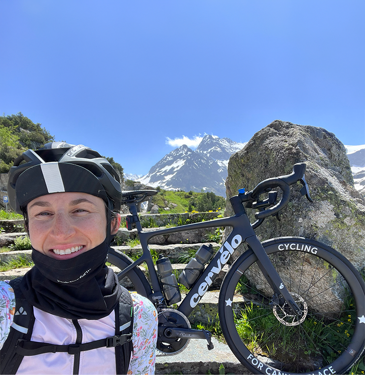 A white woman with a bright smile takes a selfie, wearing a black cycling helmet and a pink and floral-patterned cycling jersey. A black neck gaiter covers her chin and neck. Behind her, a sleek black Cervélo brand road bike with ‘Cycling for Canuck Place’ written on the wheel leans against a large rock. The background features a mountain landscape with snow-capped peaks, lush green hills, and a clear blue sky.