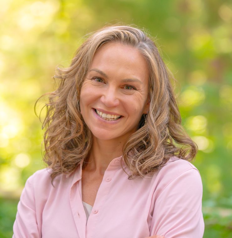 A white woman with shoulder-length curly blonde hair smiles warmly at the camera. She is wearing a light pink button-up shirt with sleeves rolled up to her elbows and has a white smartwatch and colorful beaded bracelets on her wrist. Her arms are crossed in a relaxed posture. The background consists of blurred green foliage.