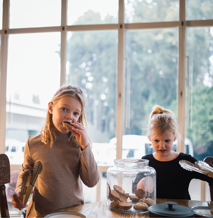 Two young girls with blonde hair stand at a wooden table in a dining area. The girl on the left has shoulder-length, slightly wavy hair and wears a long-sleeved taupe sweater. She holds a pair of silver tongs in her right hand while taking a bit of a cookie with her left, looking at the camera with a smiling expression. The girl on the right has her hair in a high ponytail and wears a black long-sleeved top. She looks at the cookies with an excited expression, holding a pair of silver tongs in her left hand. A large glass jar filled with homemade cookies sits between them.The background is blurred but shows trees and parked vehicles.