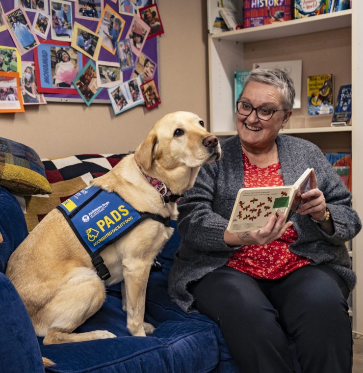 An older white woman with short gray hair, glasses, and a warm smile sits on a blue couch, holding a small book with an illustration of butterflies. She wears a red floral blouse under a grey cardigan and is engaged in reading. Beside her, a golden Labrador wearing a blue ‘PADS Accredited Facility Dog’ vest sits, gazing at the camera. The setting is a room with a bulletin board filled with colorful photos and a bookshelf stocked with children’s books.