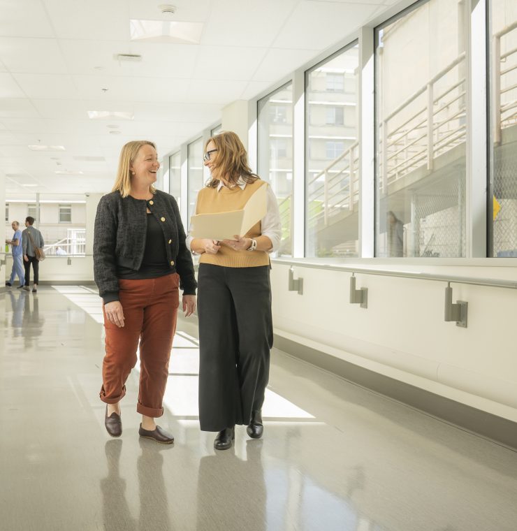 Two women walk down a brightly lit hallway while looking over papaers